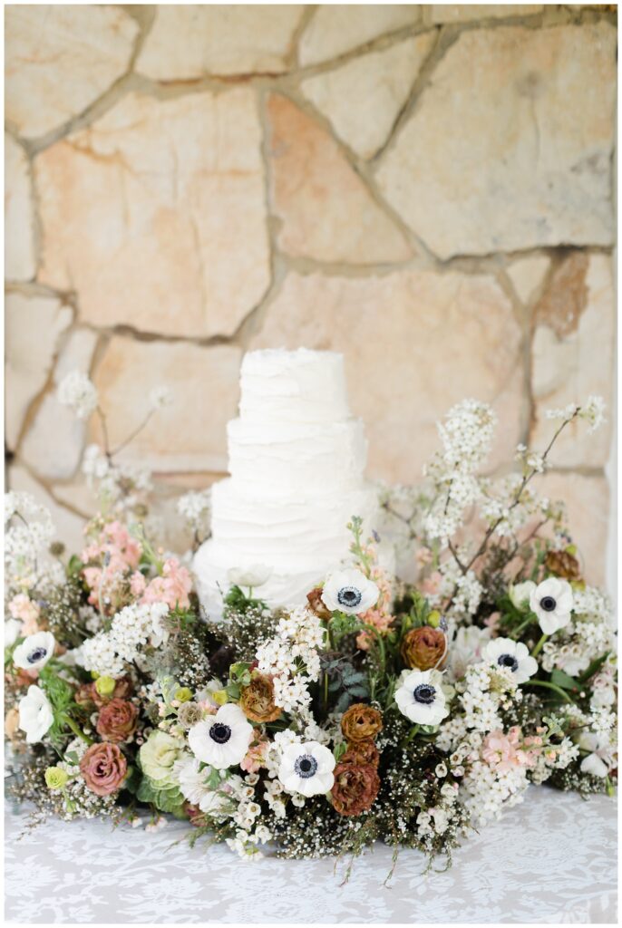 white four tiered wedding cake against a stone wall with flowers 
