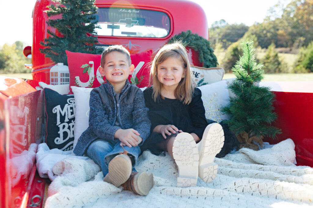 two kids sitting in the back of a red truck with Christmas decorations 