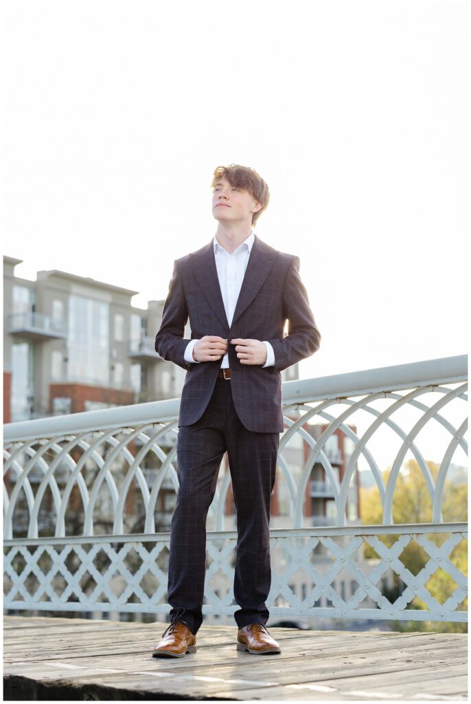 senior boy buttoning his blue jacket while posing at sunset on the Walnut Street Bridge