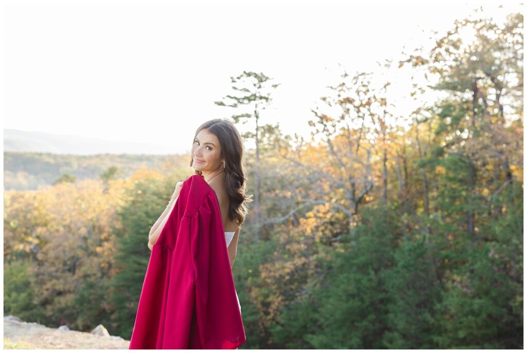 senior holding her red gown over her shoulder with mountains and trees in the background