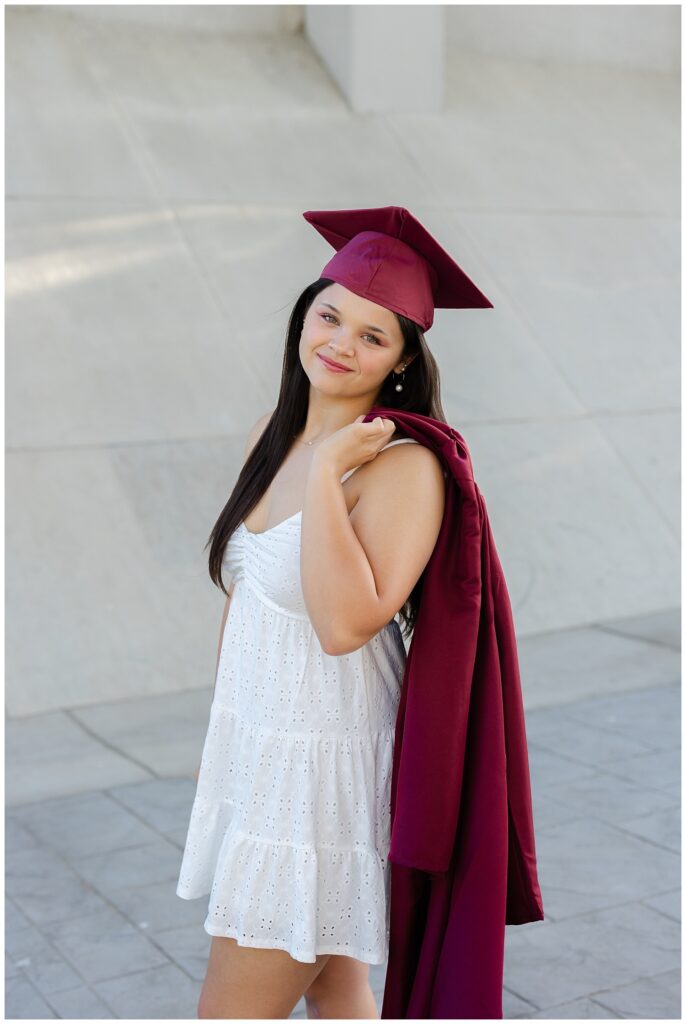 senior girl wearing a short white dress while holding a graduation gown over shoulder
