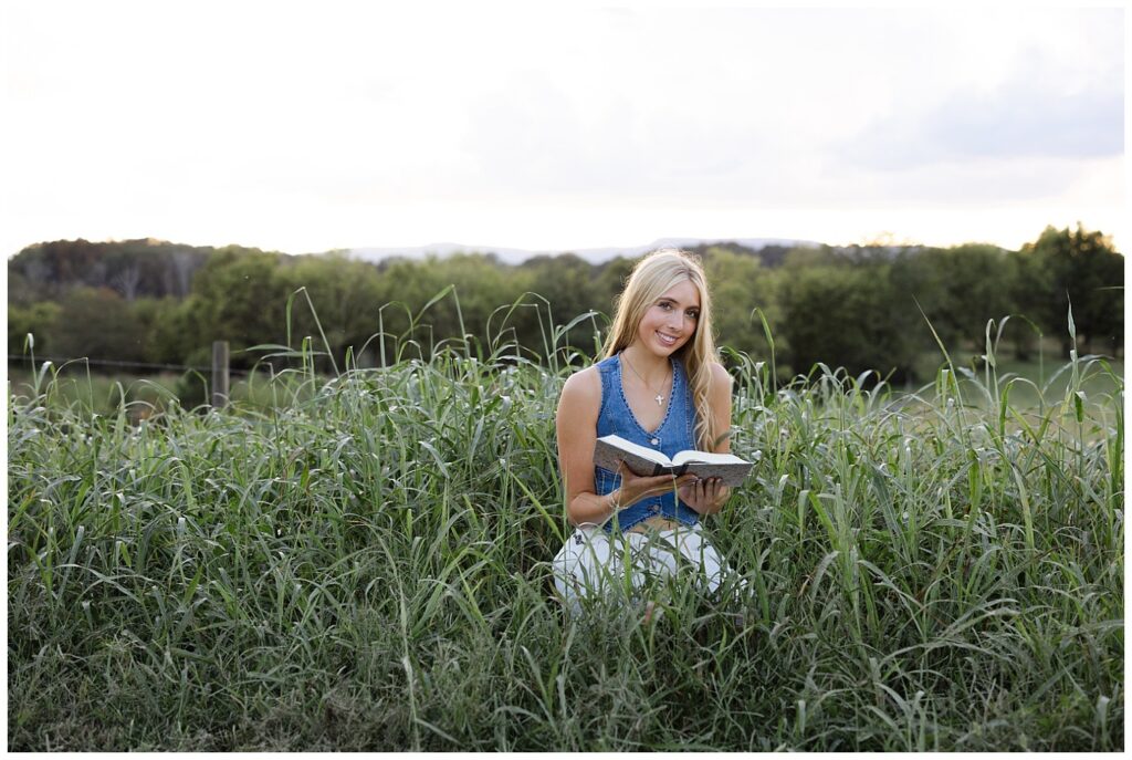 Chattanooga high school girl holding her Bible, smiling and bending down in a field on her family farm