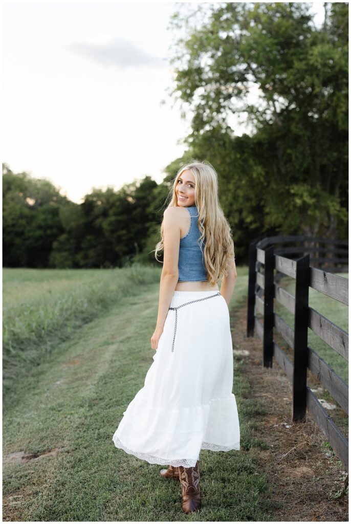 senior girl in Chattanooga looking back near a fence wearing a jean vest and white skirt and cowboy boots